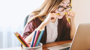 woman biting pencil while sitting on chair in front of computer during daytime