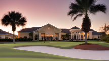 A beautifully designed golf course clubhouse surrounded by lush greenery and palm trees, set against a stunning sunset sky.