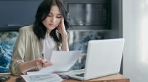 Focused young woman reviewing financial documents with a laptop in a home office setting.