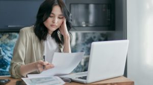 Focused young woman reviewing financial documents with a laptop in a home office setting.