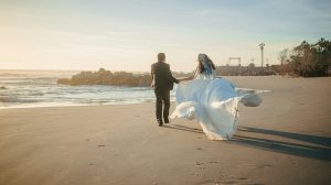 A bride and groom walk on a beach at sunset