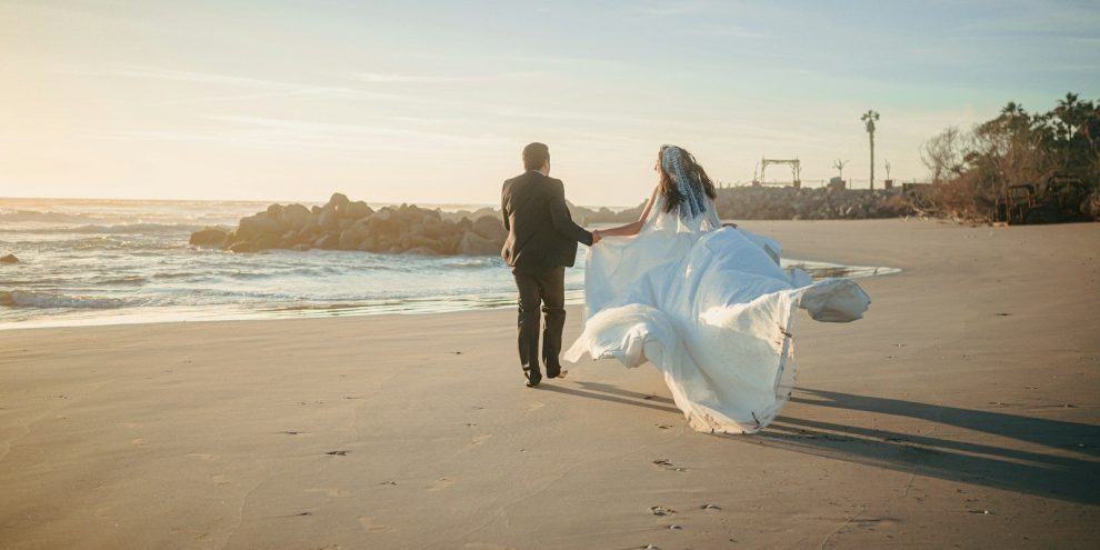 A bride and groom walk on a beach at sunset